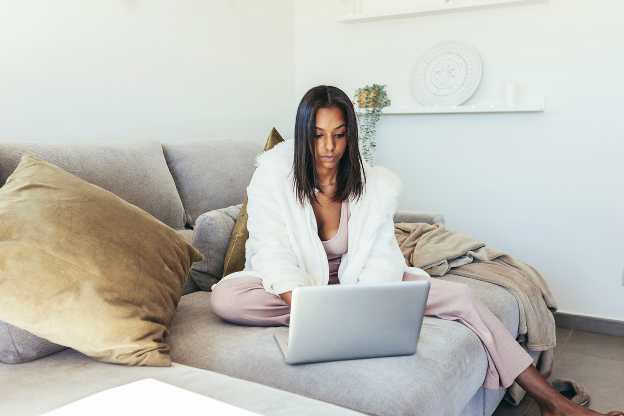 Young woman using her laptop on her couch with blanket and pillows.